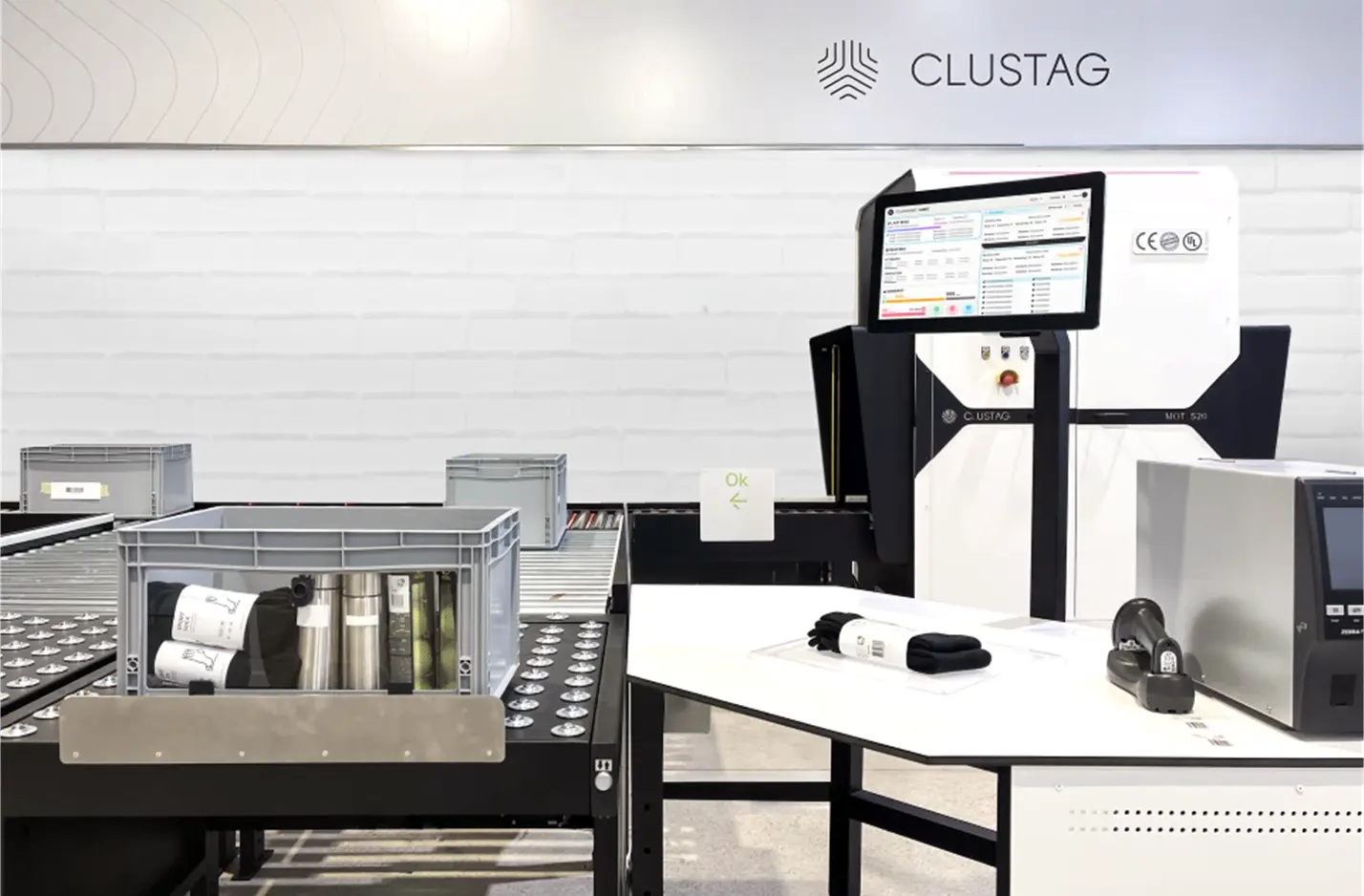 Una estación de auditoría RFID de Clustag instalada en un entorno de almacén moderno, con una cinta transportadora que lleva cubetas de plástico con artículos etiquetados, un puesto de trabajo con una gran pantalla táctil que muestra el software Zentup Workflow y varios dispositivos RFID y impresoras dispuestos sobre una mesa blanca