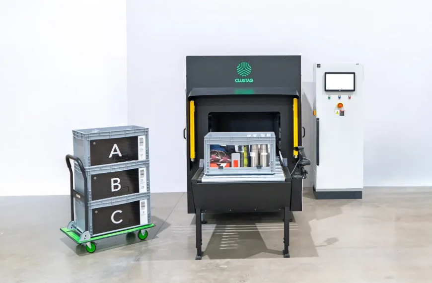 Un contenedor gris que contiene varios artículos, incluyendo unas gafas de sol, una caja de Ray‑Ban, prendas y botellas de acero inoxidable, se encuentra sobre la cinta transportadora dentro de un Clustag RFID Tunnel S24. A la izquierda, se ven tres cajas apiladas etiquetadas como A, B y C.